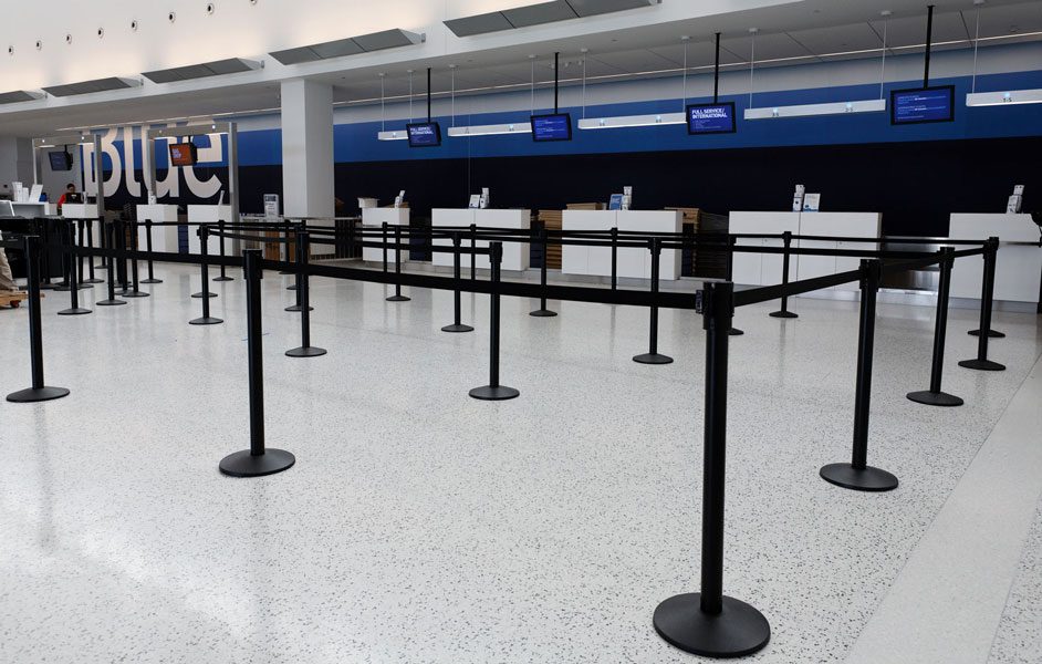 An empty airport check-in area features a JetBlue counter in the background, complemented by several rows of Visiontron retractable barriers guiding the queue on a speckled floor. Overhead, digital displays provide flight information, embodying the 6 Steps to a Great Queue Experience.