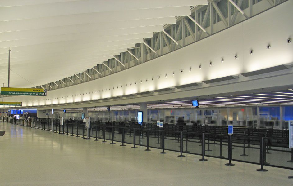 An empty airport security checkpoint area features rows of Visiontron stanchions, ensuring a great queue experience. The high ceiling showcases architectural details, with digital screens mounted above and a yellow sign visible in the background.