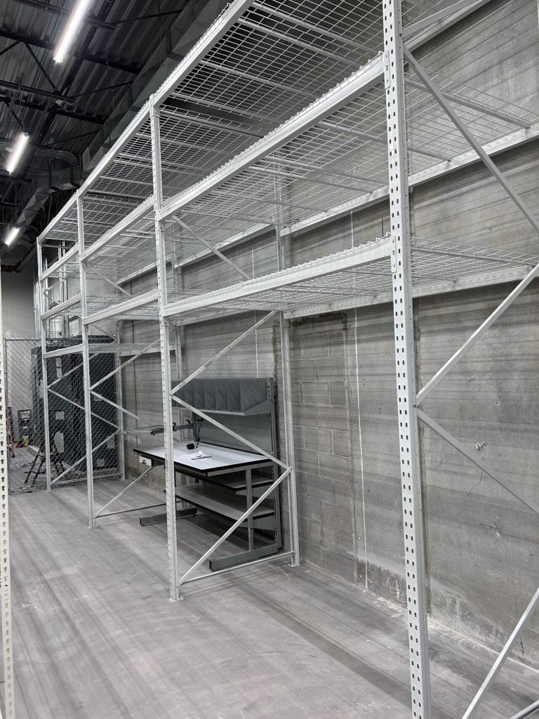A large, empty bulk rack shelving unit stands in a warehouse with a concrete floor and walls. Beneath it, theres a countertop with industrial equipment. The area is well-lit with overhead lights and partially enclosed by a metal fence.