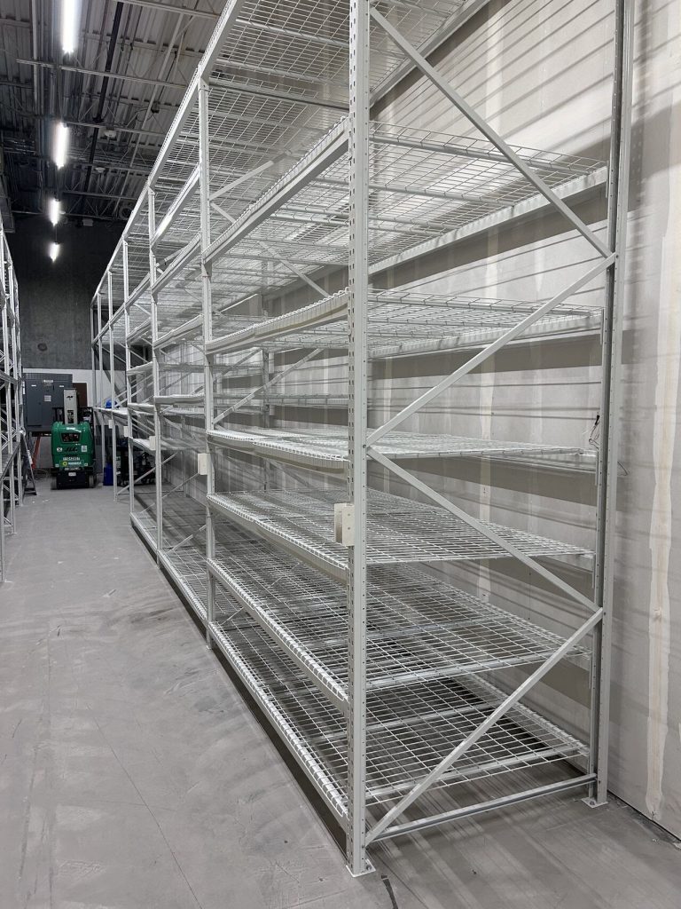 A large metal bulk rack shelving unit with multiple empty wire shelves stands in a spacious warehouse with concrete floors and high ceilings. Bright fluorescent lights illuminate the area, and machinery is visible in the background.