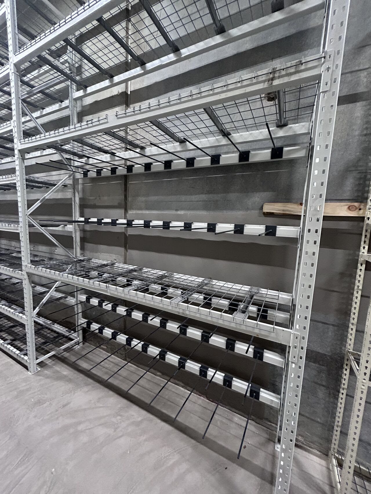 Empty metal shelving units, resembling bulk rack shelving, stand in a warehouse with a concrete floor and gray walls. The shelves have a grid-like structure and appear to be industrial storage racks.