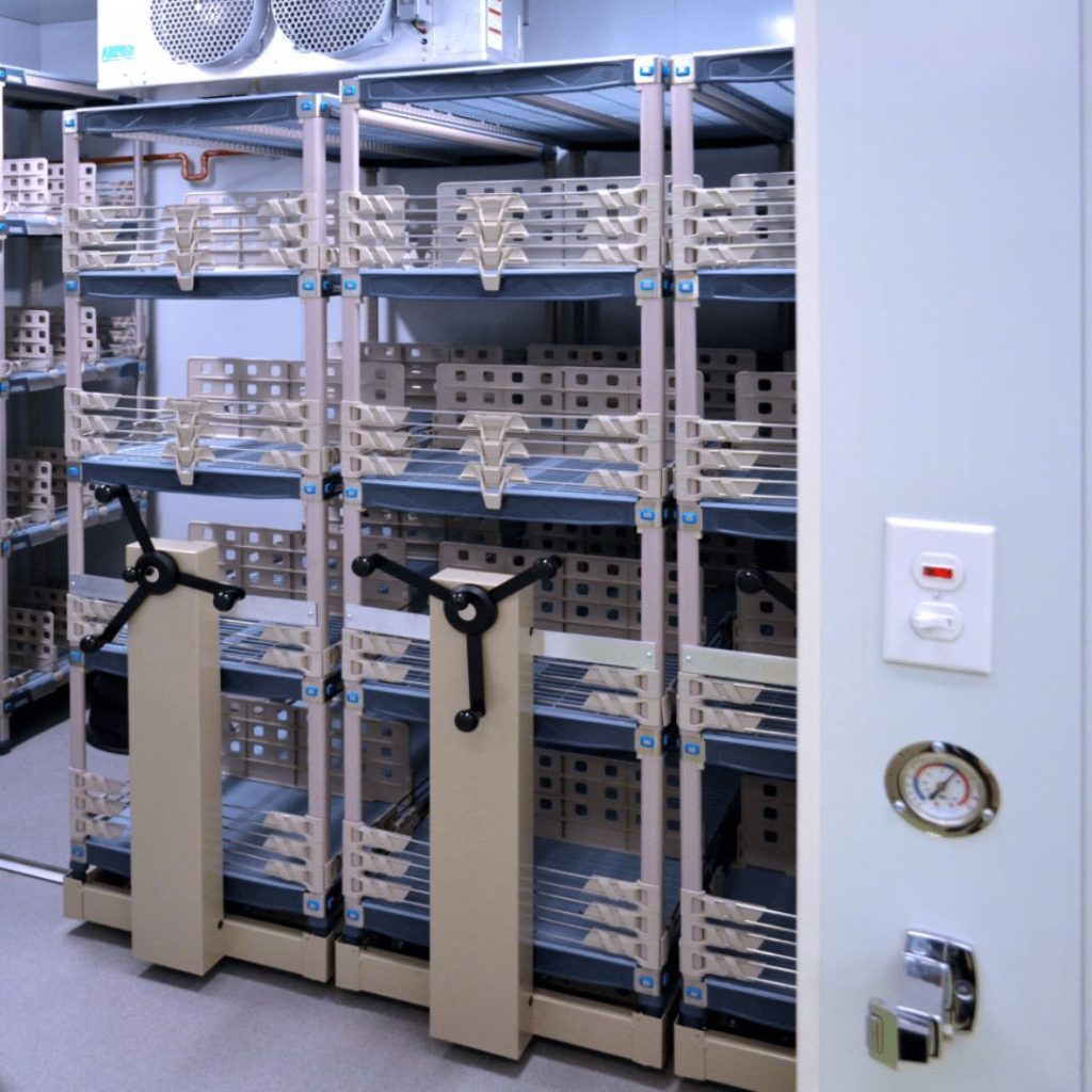 The room, an epitome of Agricultural Storage Solutions, features mobile shelving units filled with stacked trays and brick-like packages. Hand cranks adjust the shelves positions. A thermostat and control panel on the door ensure it remains organized and climate-controlled.