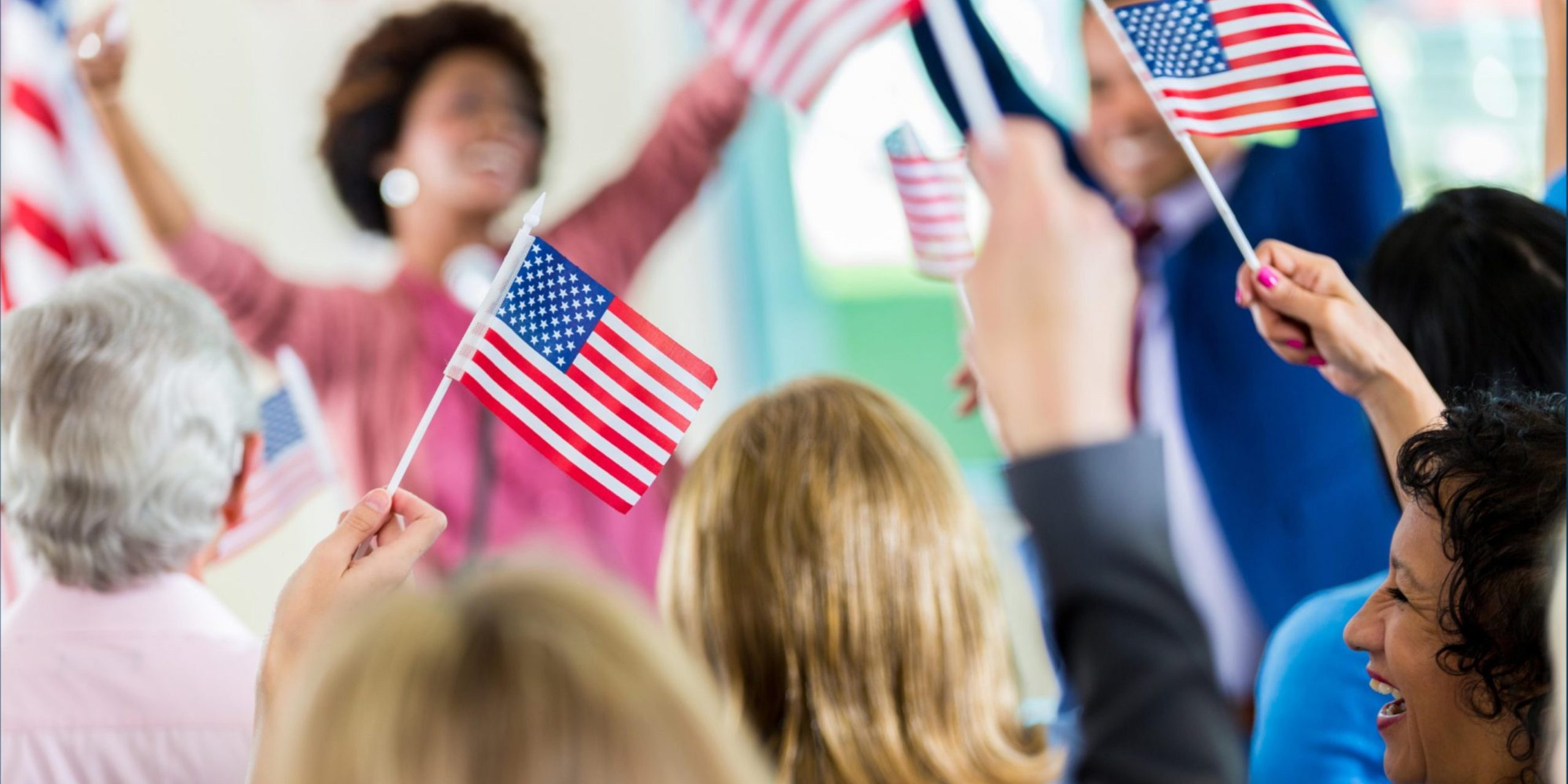 A diverse group of people enthusiastically wave small American flags, creating a vibrant scene reminiscent of crowd control at political rallies. The atmosphere is energetic and celebratory, with individuals smiling and raising their hands, emphasizing the flags patriotic unity.