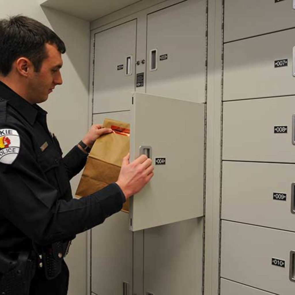 A law enforcement officer is placing a brown paper bag into a locker labeled 004 within a row of secure evidence lockers. The numbered lockers have handles for access. Dressed in full uniform, the officer remains focused on the task at hand.