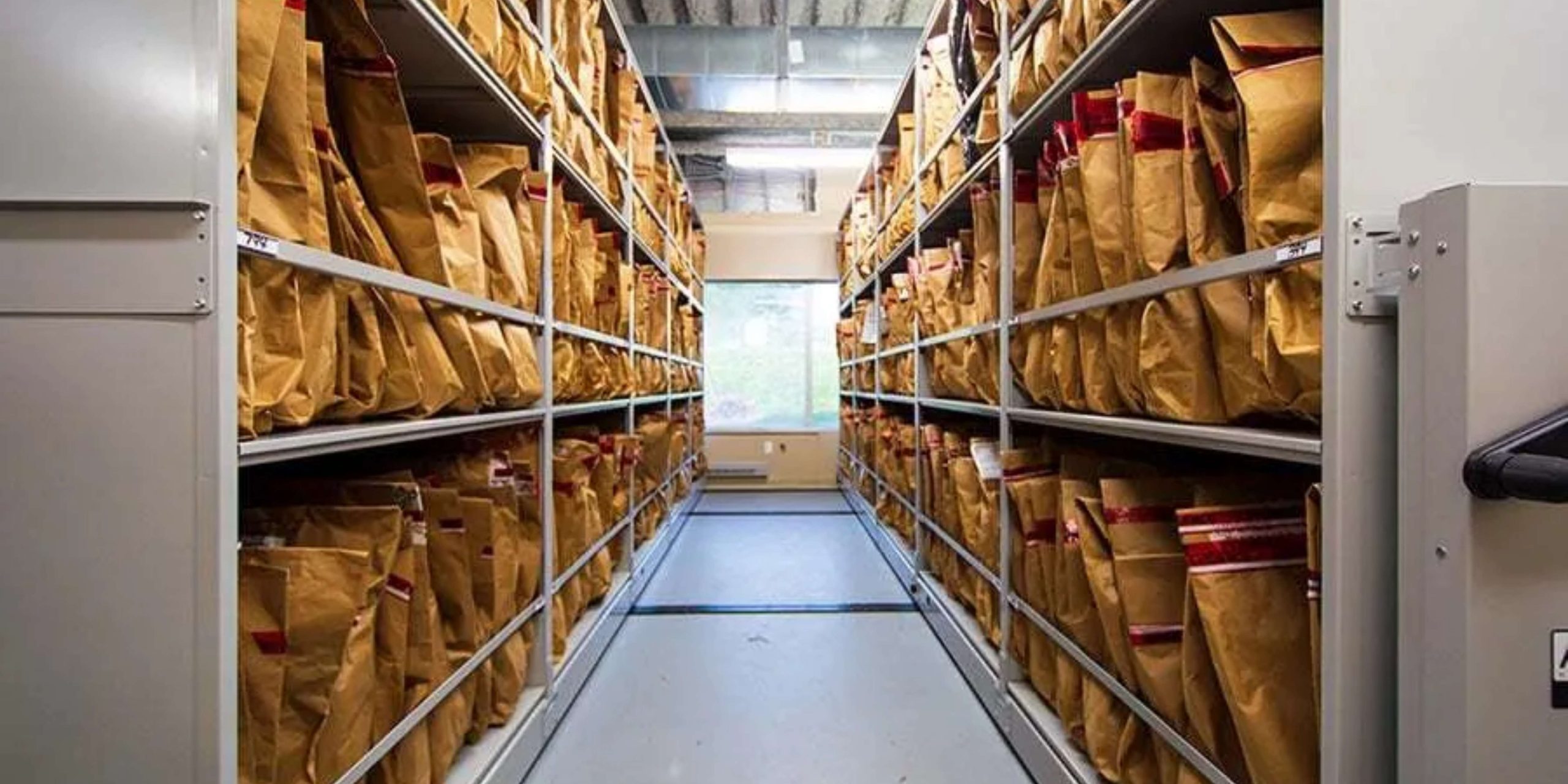 The evidence storage room design features shelves lined with brown paper bags, each sealed with red tape. A narrow walkway runs between the shelves, while natural light filters through a window in the background, highlighting the meticulously organized rows.