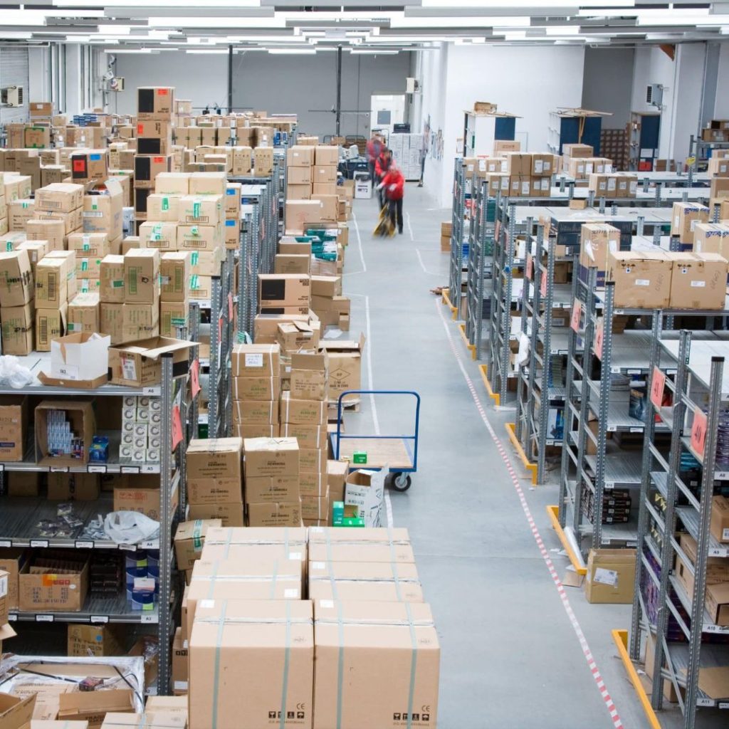 In a spacious warehouse, stacks of cardboard boxes and metal shelving units line the aisles. A person in red swiftly pushes a blue cart under the bright glow of fluorescent lights, ensuring every corner is as organized as a department in law enforcement.