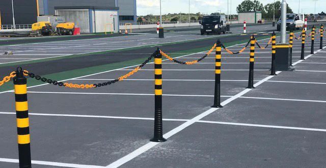 A parking lot with yellow and black posts, designed to enhance workplace safety, uses chains to separate vehicles from a pedestrian pathway. A black SUV is parked in the distance near a building. The sky is clear, and the ground is marked with white lines and green sections.