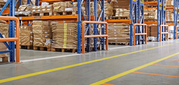 The warehouse interior features tall blue and orange metal shelving units, a vital tool for organization and safety, holding packages wrapped in brown paper. Yellow and orange safety lines mark the floors, while sturdy rails protect the shelves in this well-organized, clean space.