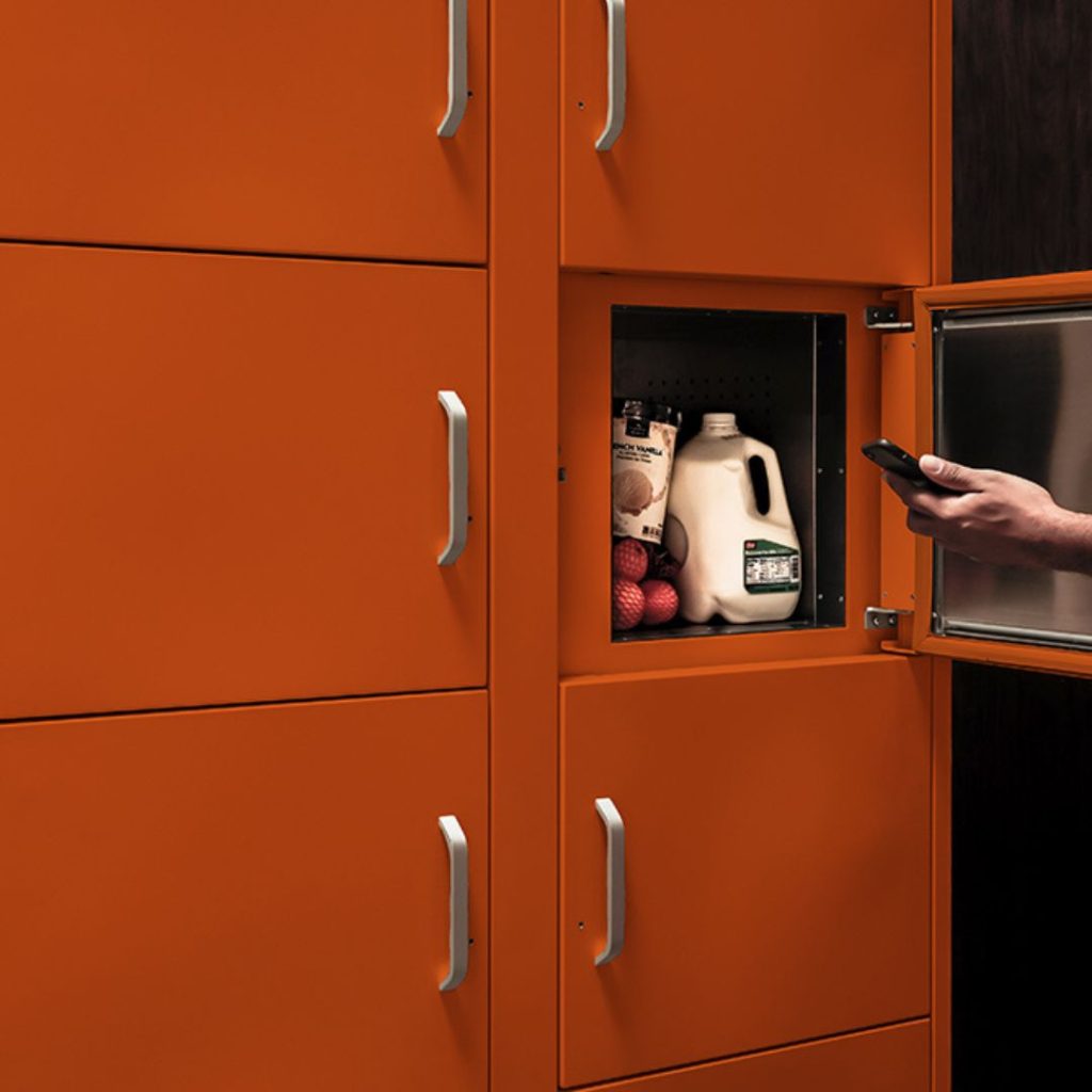 A person holding a phone stands next to modern orange kitchen cabinets, showcasing the seamless integration of property management equipment with cleverly organized spaces. One open cabinet reveals milk, apples, and coffee.