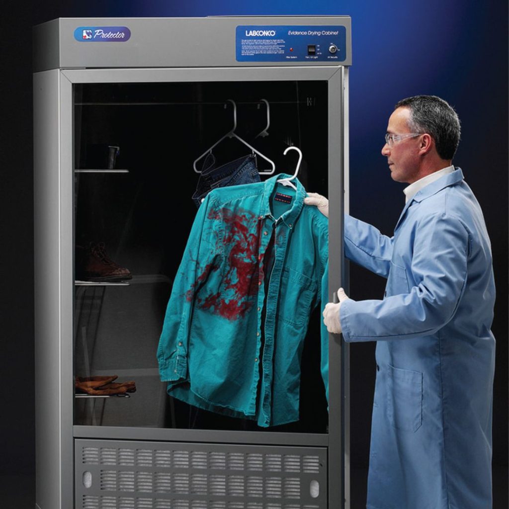A man in a lab coat examines a drying cabinet in the law enforcement department, revealing a green shirt with visible stains. Inside are also jeans and shoes. The scene suggests a meticulous investigation process amidst a scientific environment.