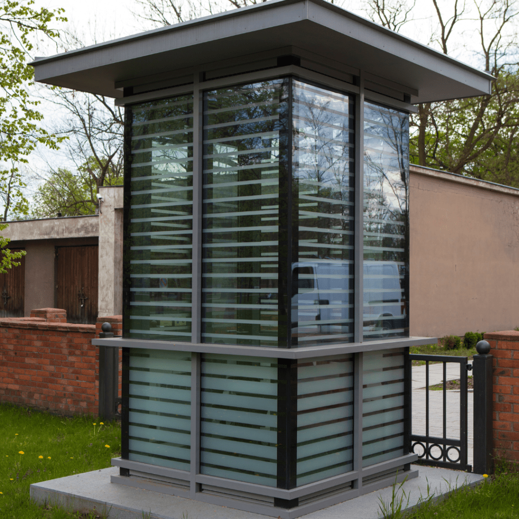 A modern glass security booth with horizontal frosted and clear panels stands next to a brick wall and gate, nestled amidst lush greenery. This government department installation features a flat roof and concrete base, blending seamlessly into its natural surroundings.
