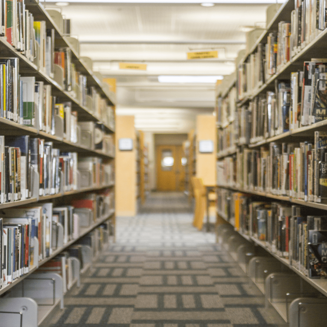 A library aisle with tall shelves brimming with educational books on both sides, leading to a well-lit nook with a wooden door at the end. The floor is carpeted in a geometric pattern, and the ceiling features fluorescent lights.