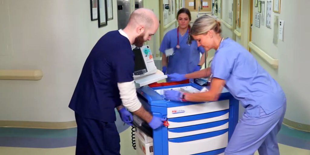 Two healthcare workers in scrubs, a man and a woman, collaborate with a Lifeline Crash Cart in a hospital corridor. Both are wearing gloves and focusing on the equipment, while another nurse walks in the background.