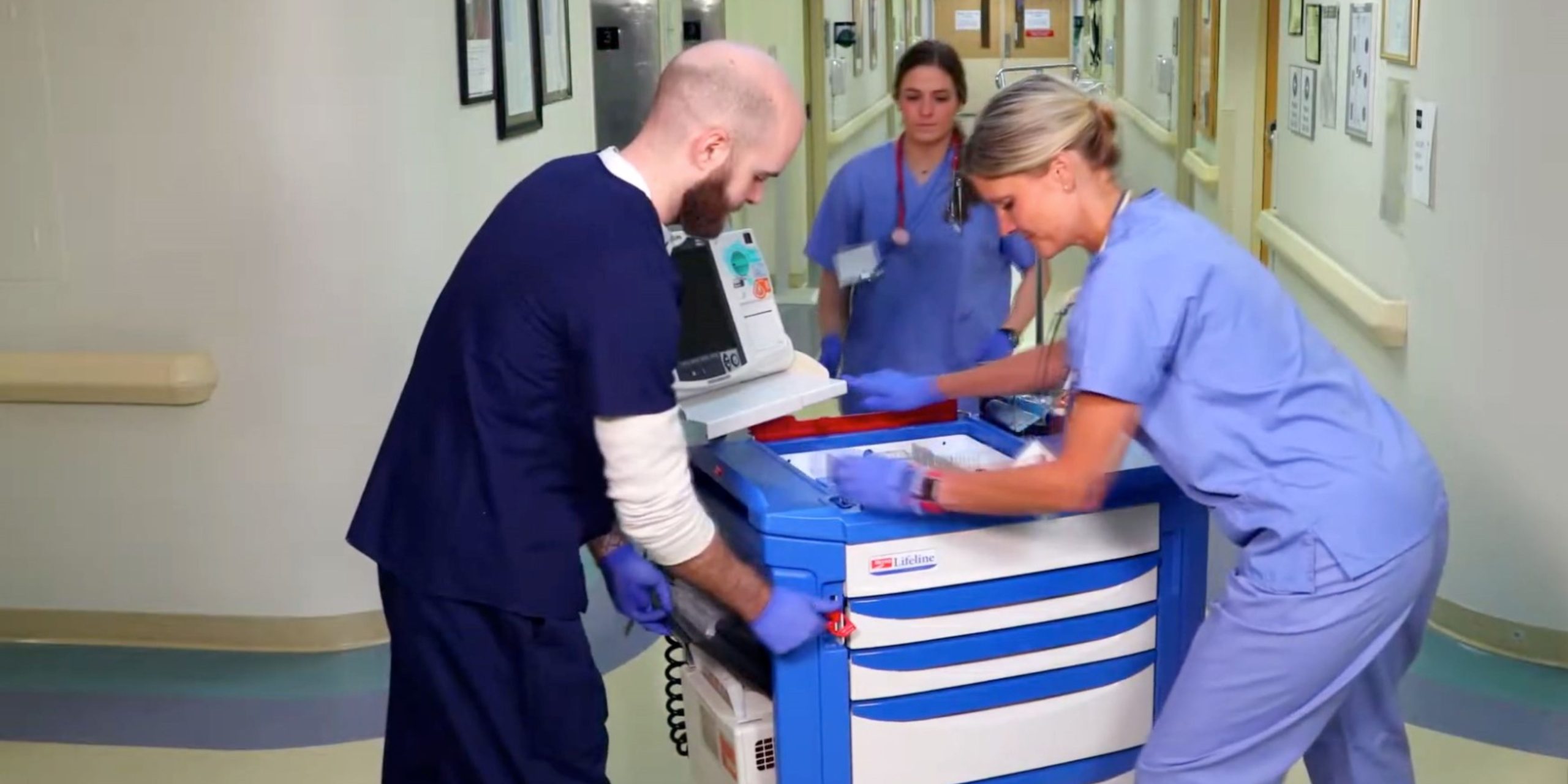 Two healthcare workers in scrubs, a man and a woman, collaborate with a Lifeline Crash Cart in a hospital corridor. Both are wearing gloves and focusing on the equipment, while another nurse walks in the background.