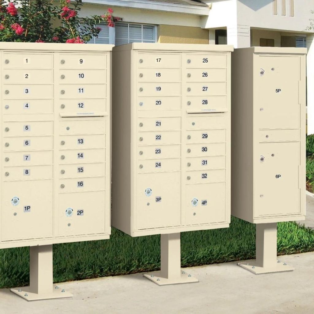 Three outdoor community mailboxes, essential pieces of property management equipment, stand on a concrete sidewalk. The background reveals green grass, shrubs with red flowers, and part of a cream-colored building.