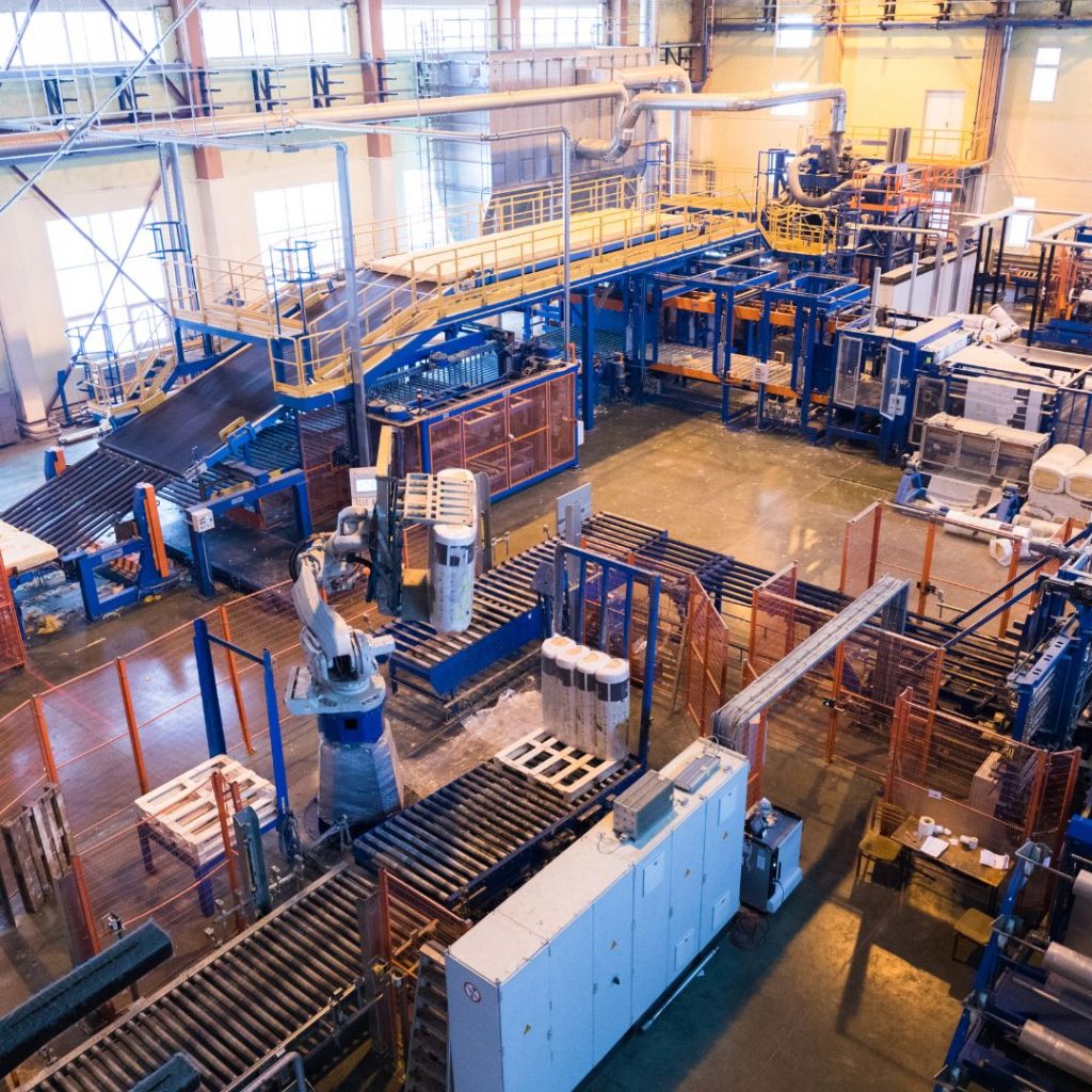 An industrial factory with machinery and conveyor belts hums with activity in the Warehouse Department. A robotic arm operates amidst various equipment, under a metallic structure with yellow railings. The shiny floor reflects the glow of overhead lights, creating a dynamic workspace.