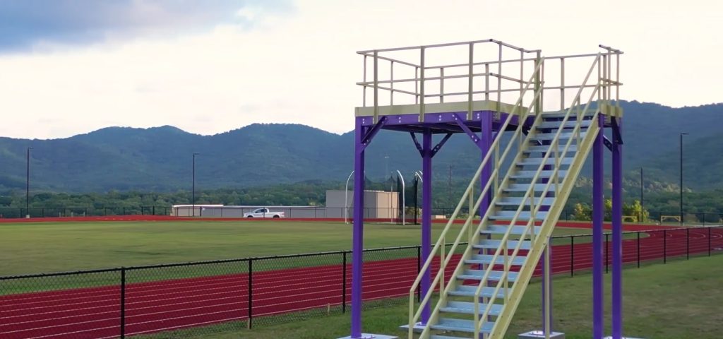 A grassy field with a red track is bordered by a black fence. A Custom Marching Band Platform stands tall in the foreground, its metal structure boasting sturdy stairs. In the background, mountains rise majestically under a partly cloudy sky.