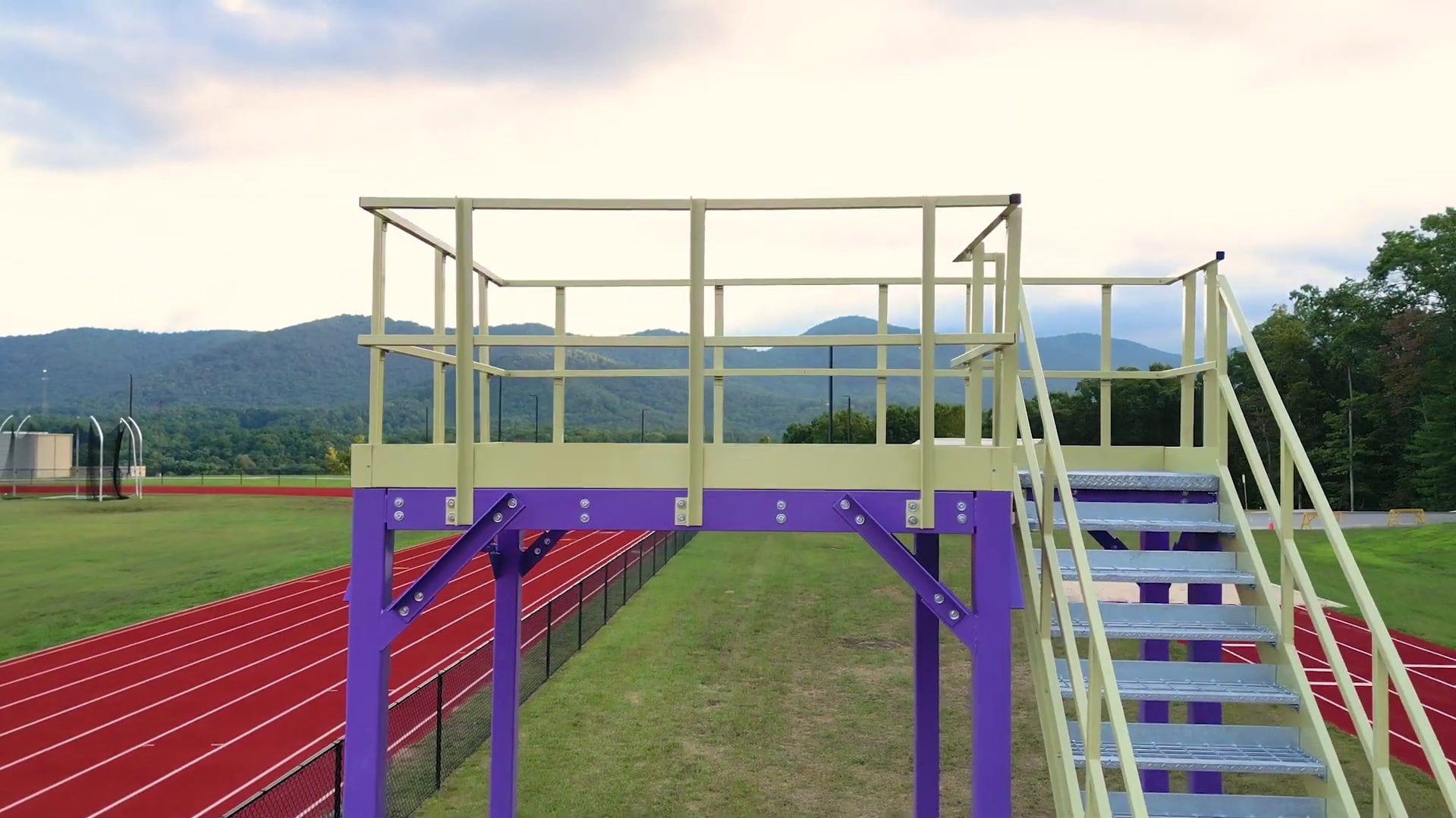 A custom marching band platform with stairs overlooks a red running track, surrounded by lush grass and trees. In the background, green mountains rise majestically under a cloudy sky, providing a picturesque setting for any performance or practice.