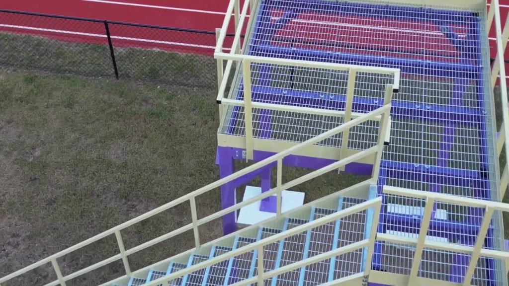 Aerial view of a Custom Marching Band Platform featuring a metal staircase painted in yellow and purple. The stairs lead to a door, enclosed by railings. Below lies a grassy area, while in the background, a red running track and black chain-link fence can be seen.