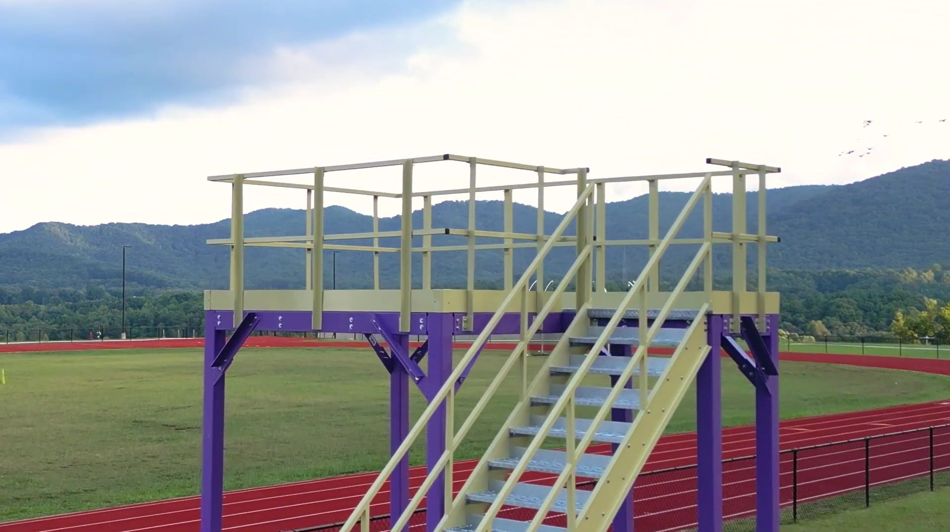 A Custom Marching Band Platform with a yellow surface and purple supports stands by a red running track. Nearby, green fields stretch towards distant hills beneath a partly cloudy sky, creating the perfect backdrop for performances.