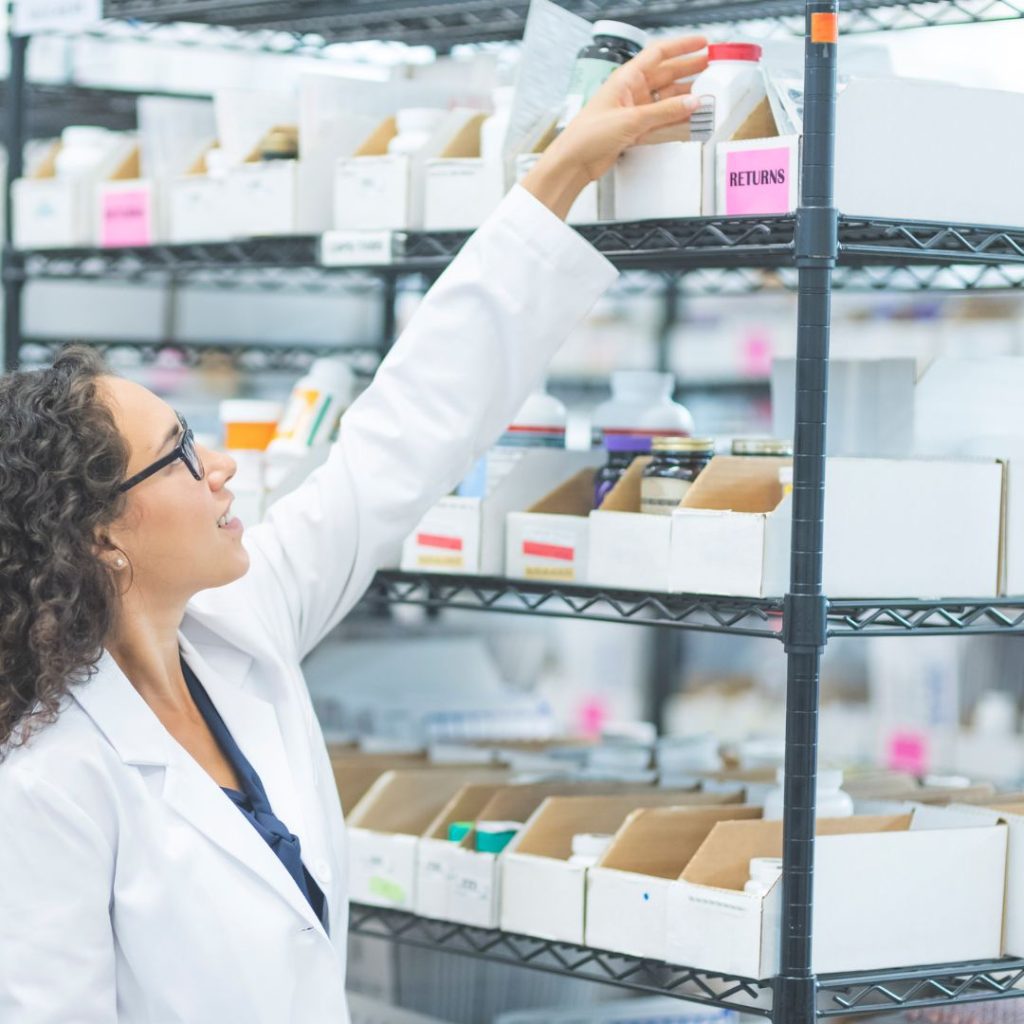 A person in a white lab coat and glasses reaches for a container on a high shelf in the healthcare storage area, filled with boxes labeled Returns. Shelves are stocked with various bottles and containers.