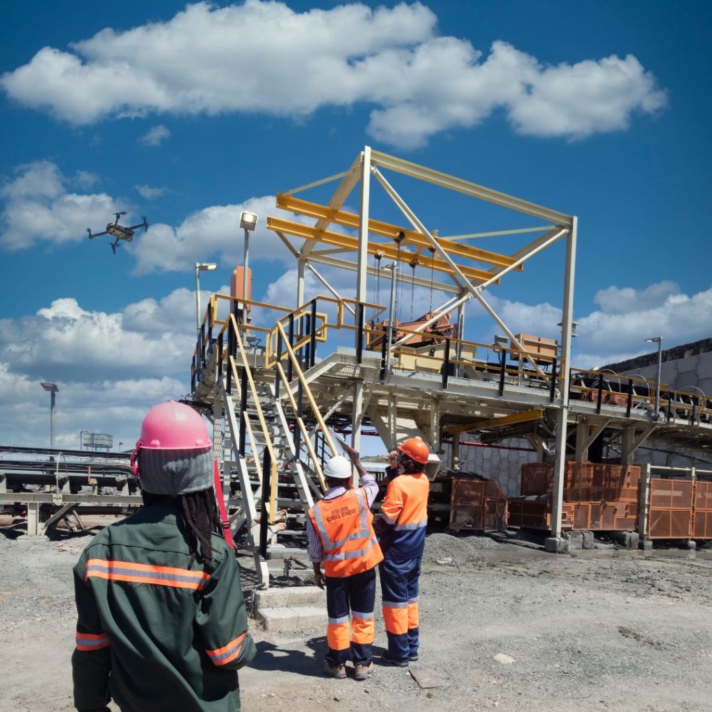 Workers in protective gear stand near industrial equipment at a bustling construction site. One observes a drone flying overhead, while the departments warehouse operations continue seamlessly. A bright blue sky with clouds forms the perfect backdrop.