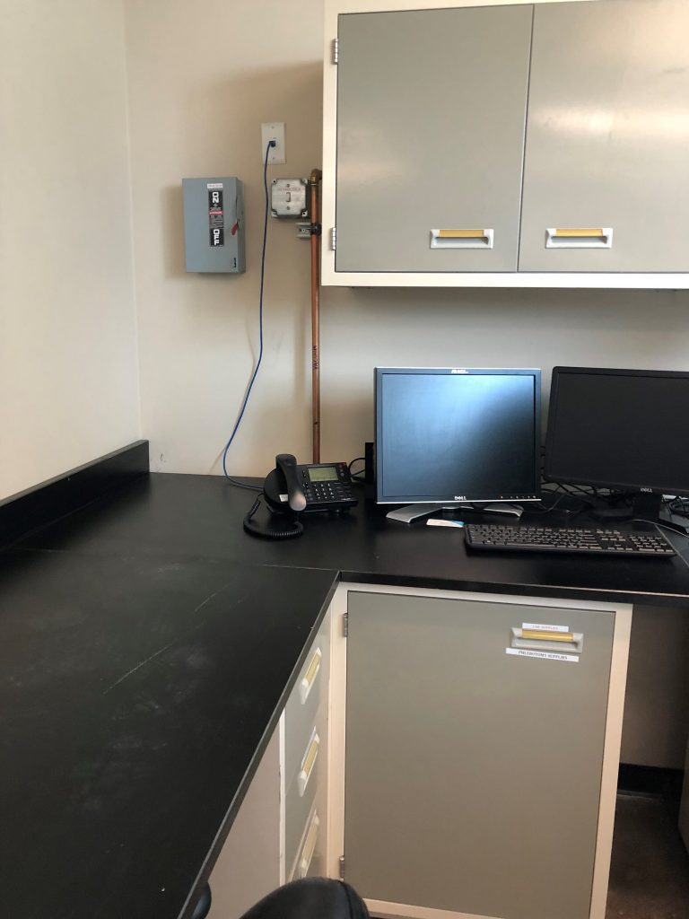 A tidy office workspace with a black countertop showcases high-quality laboratory furniture, perfect for analytical testing. It features two monitors, a keyboard, mouse, and phone. Gray cabinets with white handles are mounted above the desk, and a power box is visible on the wall.