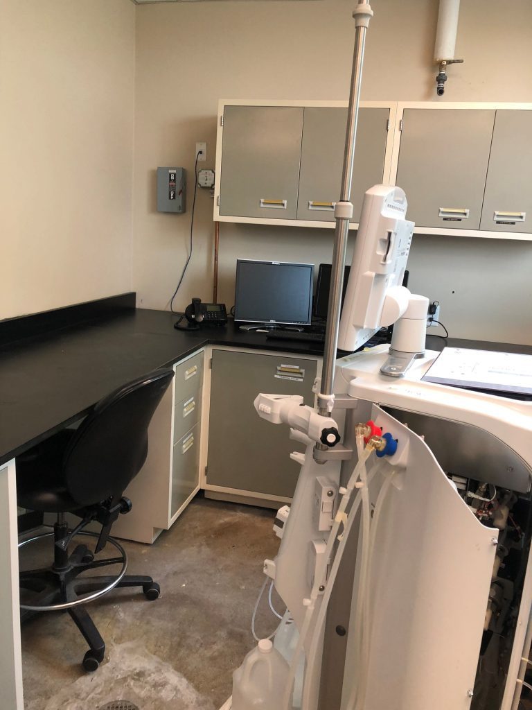 A small laboratory room features high-quality laboratory furniture for analytical testing, including a workstation with a chair, computer, phone, and cabinets. In the foreground, medical equipment with various tubes and controls enhances its sterile and organized appearance.