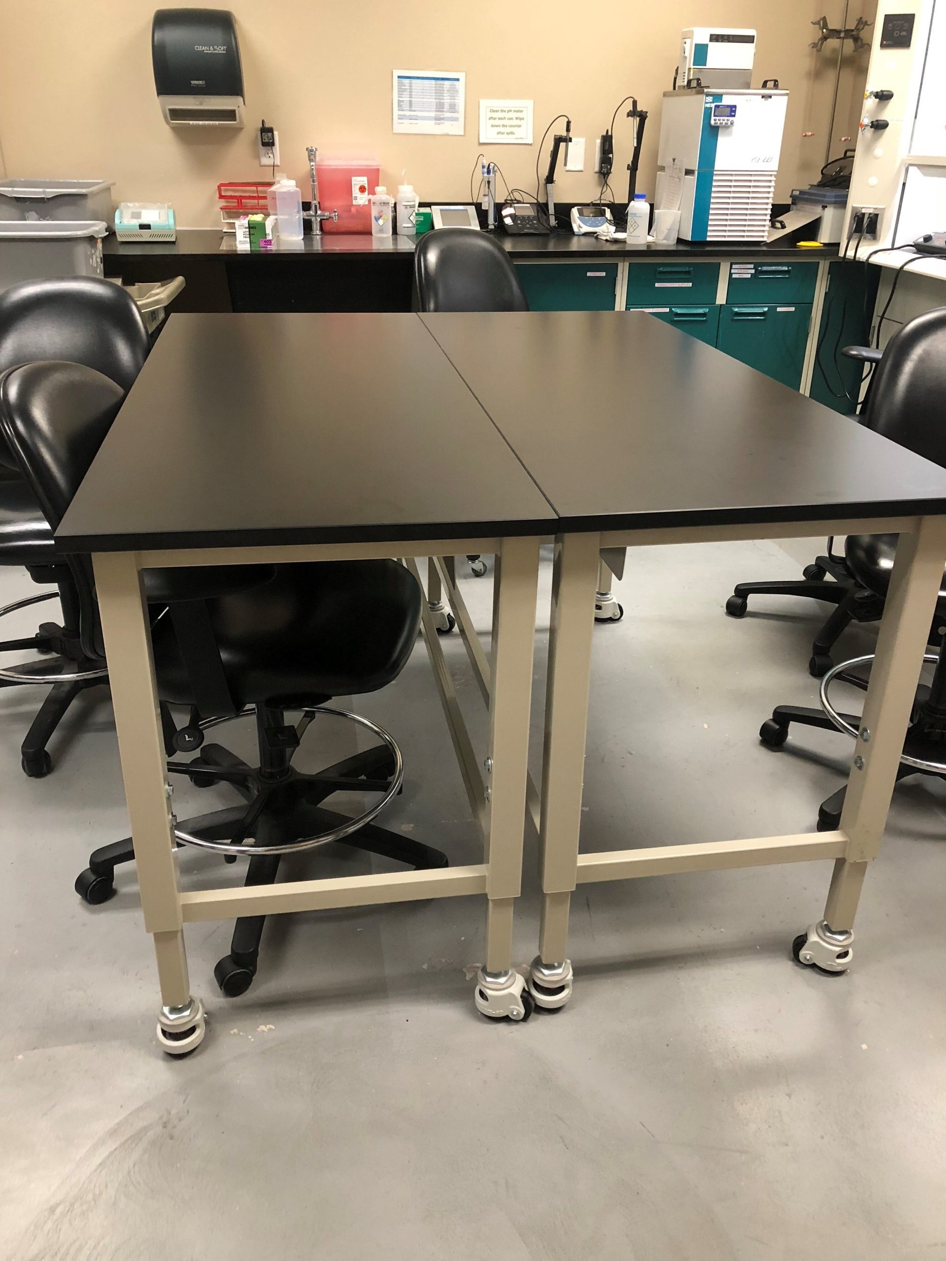 A laboratory workspace features high-quality black tables for analytical testing, pushed together and surrounded by several black swivel chairs. In the background, there are cabinets, shelves with various lab equipment, and a paper towel dispenser on the wall.