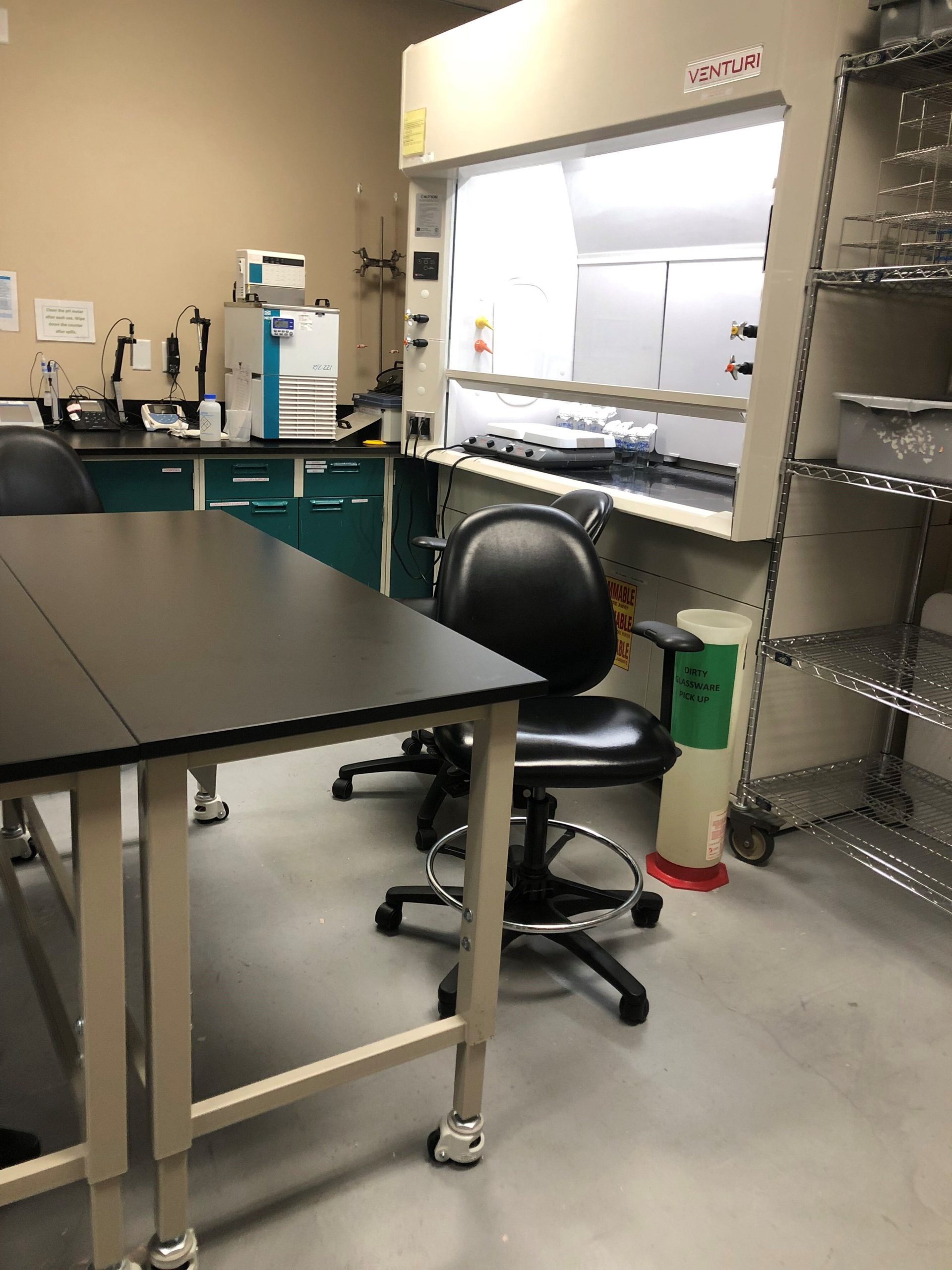 A high-quality analytical testing laboratory boasts a black tabletop, rolling chair, and ventilation hood. Shelves with storage containers line the right side, while a water purifier and lab gadgets sit on the left. The gray flooring complements the beige walls seamlessly.
