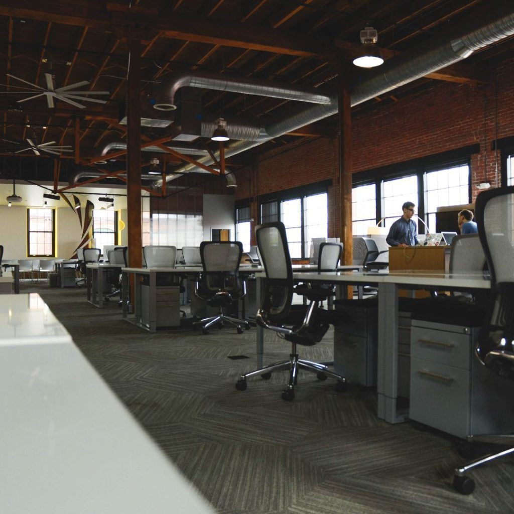 Spacious open-plan office with rows of empty workstations and office chairs on carpeted flooring. Exposed wooden beams and brick walls lend an industrial feel reminiscent of a warehouse. A few people are gathered in the background near large windows.