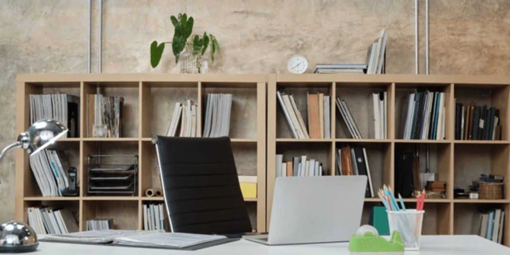 A modern office space features an empty black chair at a sleek white desk adorned with an open folder, laptop, desk lamp, and stationery holder. Behind it lies an impressive display of office shelving images on a wooden bookshelf filled with books and decor items.