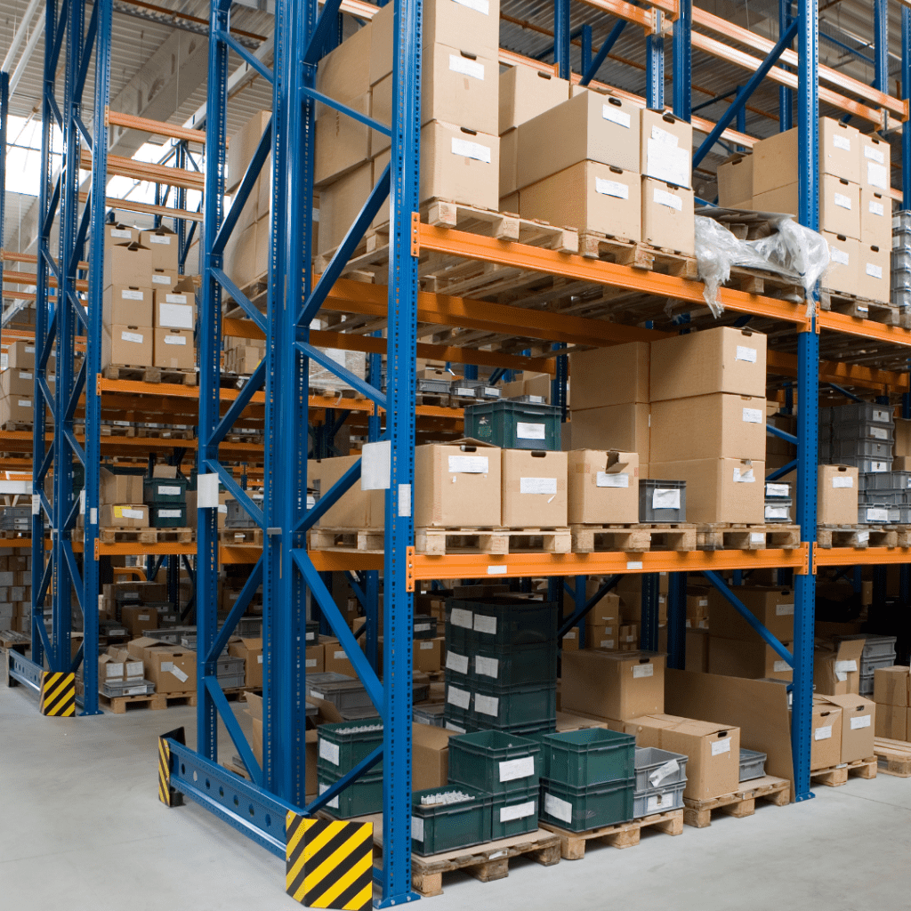 The warehouse interior features tall blue and orange shelves, resembling an organized library from the Department of Education. Various cardboard boxes and plastic containers are neatly labeled and stacked in rows. The clean floor and ample lighting enhance the orderly environment.