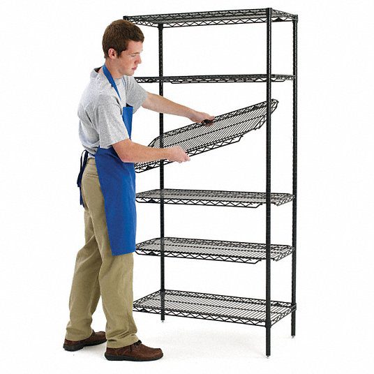 A person in a blue apron and khaki pants is assembling a black metal shelving unit, reminiscent of those found in Supermarket Solutions. They are inserting a shelf into the rack, which has four other shelves already in place. The background is plain white.