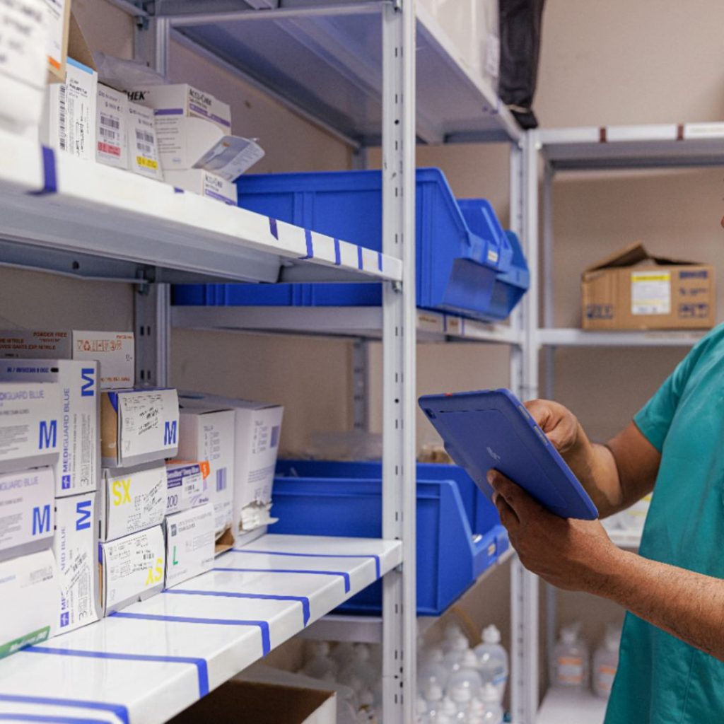 A person in a green shirt holds a clipboard, overseeing healthcare supplies neatly organized in boxes and blue bins on metal shelves in the storage room.