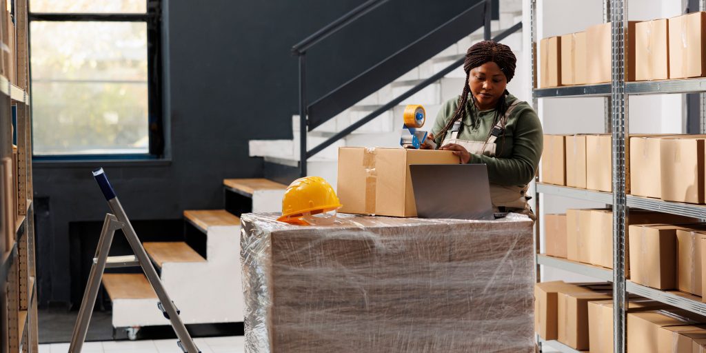 A woman tapes a cardboard box on a pallet in the warehouse, where inventory management solutions are evident. A yellow hard hat rests on the wrapped pallet, an open laptop is nearby, and a ladder stands in the foreground. A staircase and shelving units stretch into the background.