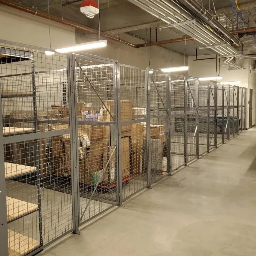 Row of wire mesh storage enclosures with shelving and cartons along a facility corridor