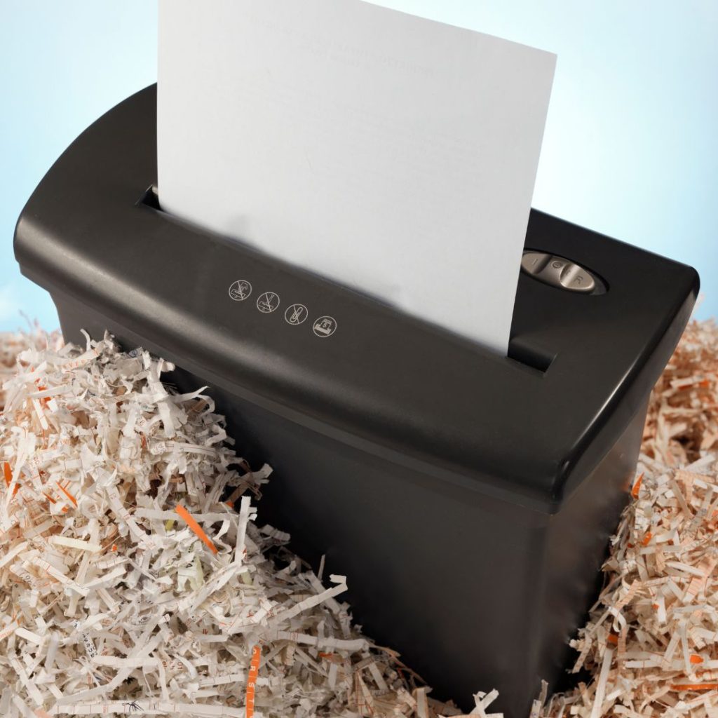 A black paper shredder efficiently shreds a piece of paper, surrounded by a pile of shredded strips. Positioned against a light blue background, this perfect office accessory ensures your department maintains confidentiality with ease and style.