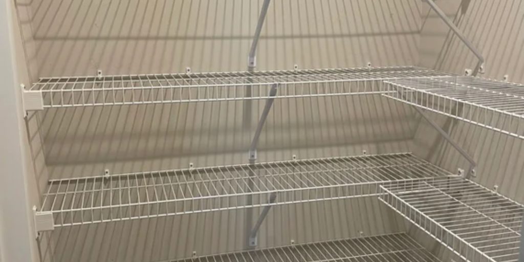 Empty white wire shelving unit with three tiers stands in a storage or closet space. The shelves, featured prominently in wire shelving images, are supported by diagonal metal bars against a plain, light-colored wall, while shadows from the shelves create an intriguing pattern.