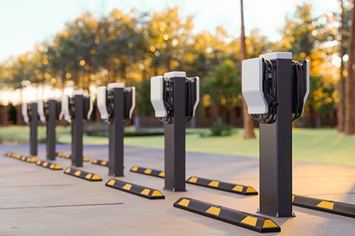 A row of electric vehicle charging stations is set in a sunny outdoor setting, ideal for workplace charging, with trees and a lawn as the backdrop. Each station features charging cables and black and yellow wheel stops in front.