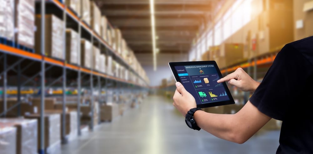In a bustling warehouse, a person uses a tablet to oversee inventory management with advanced picking and packing solutions. Shelves brimming with boxes form the backdrop as the screen showcases graphs and data on stock levels, ensuring seamless operations.
