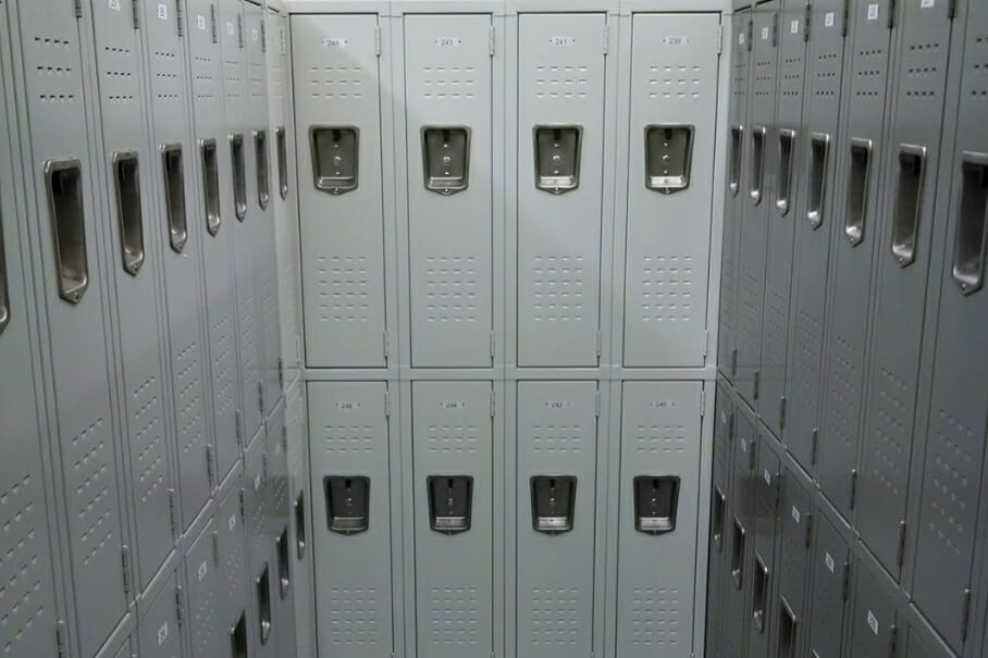 A room filled with rows of gray metal lockers, each featuring vents and a handle, reminiscent of streamlined food service shelving solutions. These lockers form an L-shape along two walls, creating a tight, enclosed space bathed in overhead lighting.