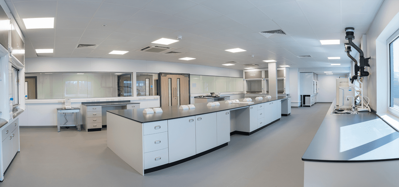 Panoramic view of a modern laboratory with sleek white counters and cabinets. The room is spacious, featuring overhead lighting and large windows on the right. An array of lab equipment gleams along the remodeled countertops designed for optimal use.