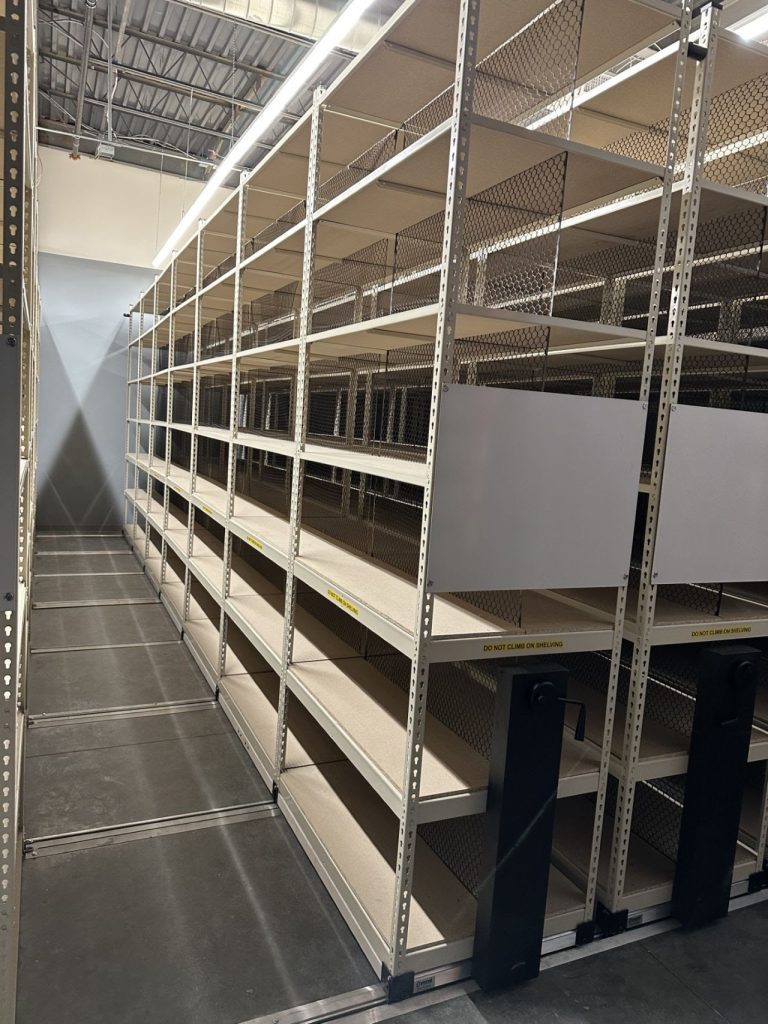 A long, empty metal shelving unit inside a storage or warehouse facility stands ready to serve as part of efficient agricultural storage solutions. The shelves are bare with mesh sides, and the area is well-lit with overhead lighting.