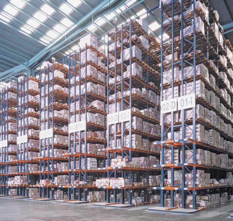 Rows of tall blue metal shelves stacked with boxes and goods in a vast warehouse with a high ceiling, optimized for efficient picking and packing solutions. Large signs with numbers are attached to the racks for organization, while a concrete floor spans the warehouse.