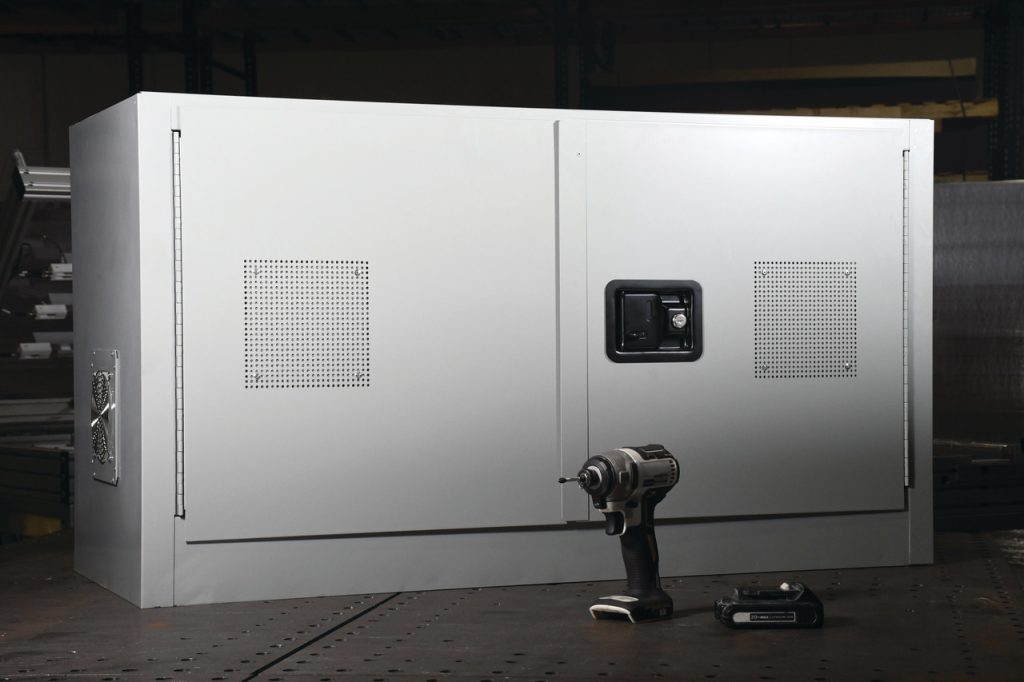 A large gray metal cabinet featuring ventilation panels and a black handle sits on a table, housing the ChargeGuard™ Containment System. A cordless drill with a battery lies in front of it, highlighting its role in lithium battery storage solutions within the dimly lit workshop.