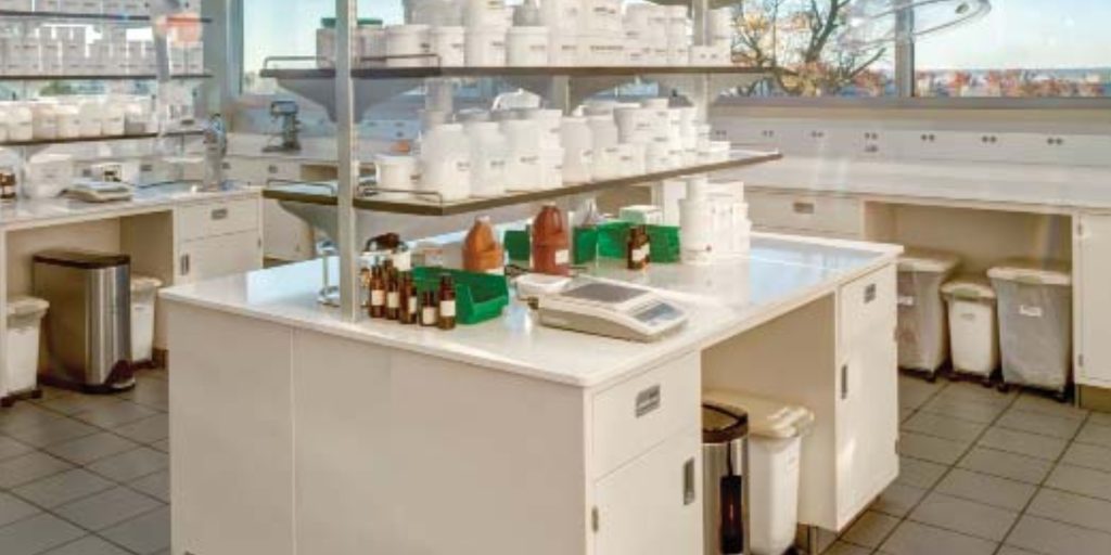 A clean, modern laboratory with a central workstation featuring various containers, bottles, and a scale equipped with custom drawer slides. Shelves above hold numerous white jars. Large windows in the background offer a view of trees and a clear sky, enhanced by electronic locking systems for security.