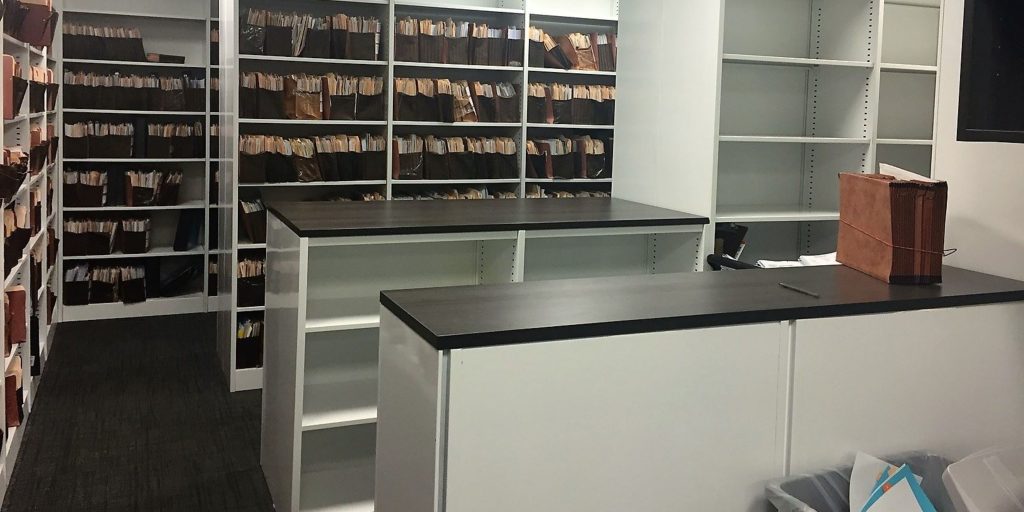 The room features two dark countertop desks and white shelves filled with organized files and folders, showcasing the efficiency of Amscor Storage Solutions. Neatly arranged, one brown file box sits on a desk, while papers are neatly placed in bins below.