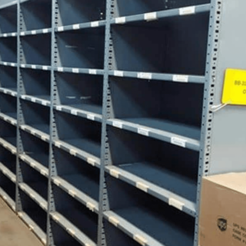 Empty metal shelving units with multiple levels stand in a room that resembles a gallery of functional design. A yellow sign peeks from the right, while a brown cardboard box sits in the bottom right corner. The shelves are labeled but lack items, creating an exhibit-like atmosphere of material handling.