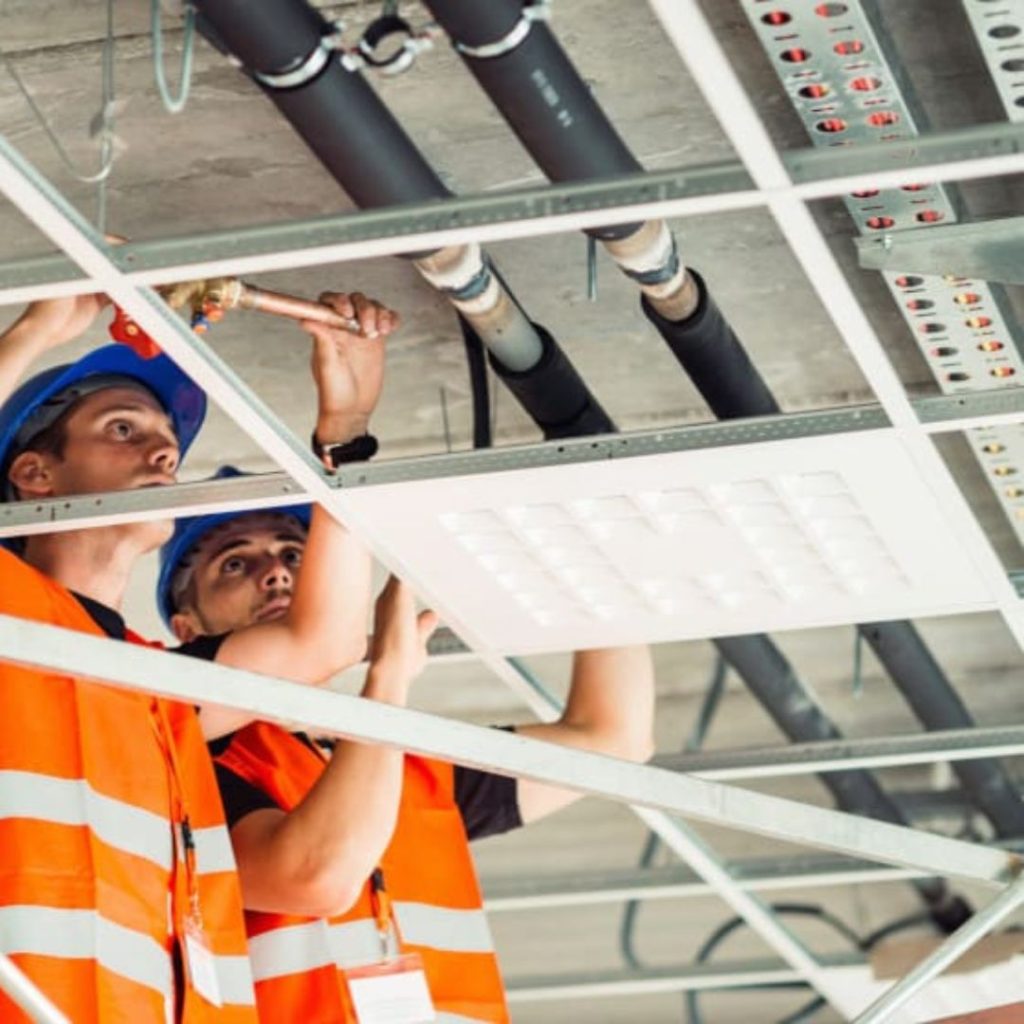 Two workers in blue helmets and orange safety vests are diligently installing or inspecting ceiling pipes and a panel light. One holds a tool while the other assists, ensuring everything is perfect for this buildings future role in the Department of Educations initiatives.